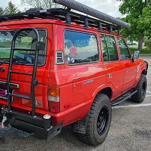 Rear-side view of a red Toyota Land Cruiser with off-road tires, a roof rack with an awning, and a ladder mounted to the back. The glossy paint and tinted windows reflect a well-maintained, adventure-ready vehicle.