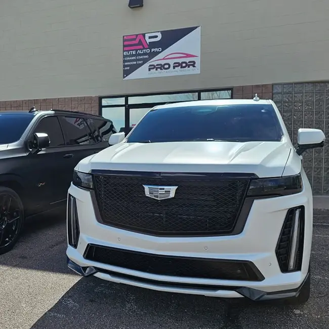 Two luxury SUVs, including a white Cadillac Escalade with blacked-out grille and trim, are parked in front of the Elite Auto Pro and Pro PDR storefront. A sign above the entrance displays both business names.