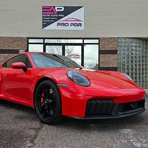 A bright red Porsche sports car with black wheels is parked in front of the Elite Auto Pro and Pro PDR shop. The building features large windows and a dual-branded sign above the entrance.