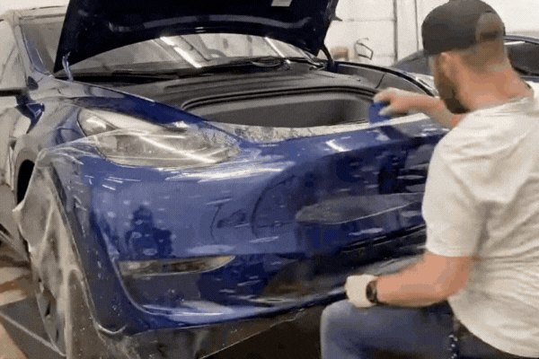 A technician applies a clear paint protection film (PPF) to the front bumper of a blue Tesla with the hood open. The specialist uses a squeegee to smooth out the film, ensuring a seamless, bubble-free finish to protect the vehicle’s paint from damage.