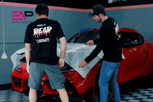 Two professional installers carefully apply a large sheet of paint protection film to the hood of a red sports car inside the Elite Auto Pro studio. Both wear black shirts with the words “WORK IS DEAD” and the shop’s logo, emphasizing expert, bubble-free installation.