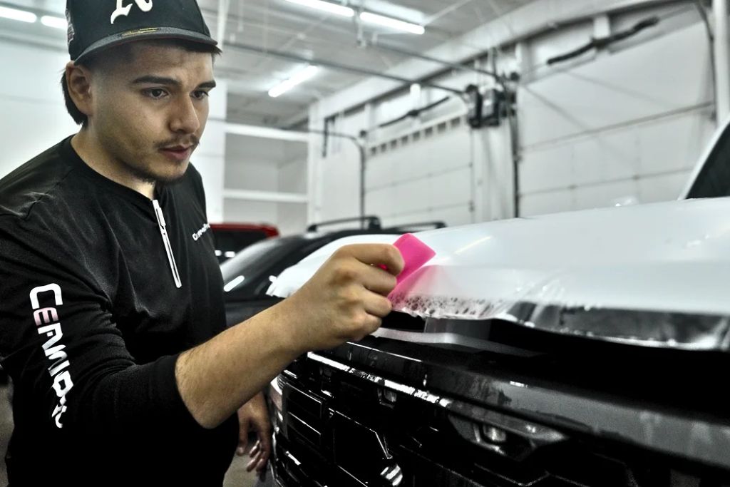 A technician in a black shirt uses a pink squeegee to carefully apply clear paint protection film to the hood of a white truck inside an auto shop. The close-up highlights the attention to detail required for a seamless, protective finish.