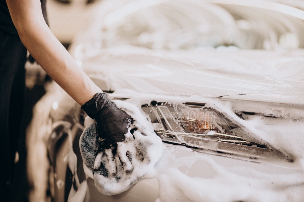A person wearing a black glove carefully hand-washes a car’s headlight and fender area with a microfiber mitt and thick, soapy foam. The image emphasizes gentle, hands-on care—one of the best practices for long-term protection of ceramic coatings and PPF.