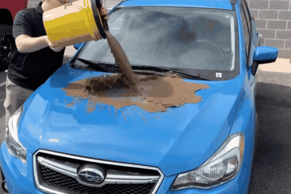 A person pours a large bucket of mud directly onto the hood of a bright blue Subaru, demonstrating the protective power of ceramic coating as the muddy water quickly slides off the surface. The scene highlights the hydrophobic and self-cleaning effects of ceramic coating in action.