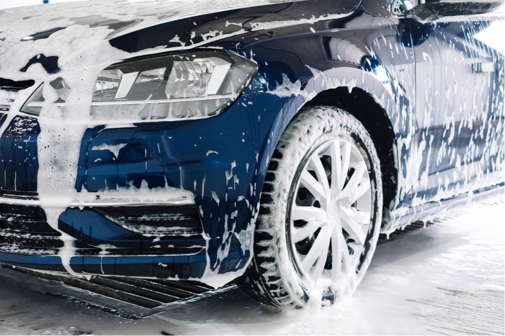Close-up of a blue car covered in soap and foam during a winter wash in Colorado Springs, with emphasis on the front bumper and wheel area where salt and grime accumulate. The image underscores the importance of regular cleaning to maintain colored PPF performance.