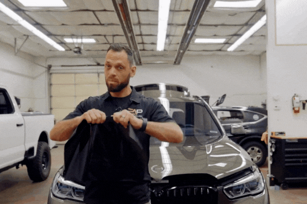 A technician at a Colorado Springs auto shop demonstrates the strength of colored paint protection film (PPF) by pulling and stretching a black sheet of film in front of a dark SUV. The workshop setting highlights the hands-on durability test of PPF over vinyl wraps.