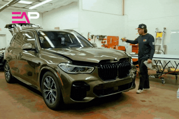 Inside Elite Auto PRO's Colorado Springs studio, a technician prepares a metallic bronze BMW SUV for colored PPF installation, surrounded by tools and equipment. The Elite Auto PRO logo appears in the corner, reinforcing the shop's professional, high-quality service.