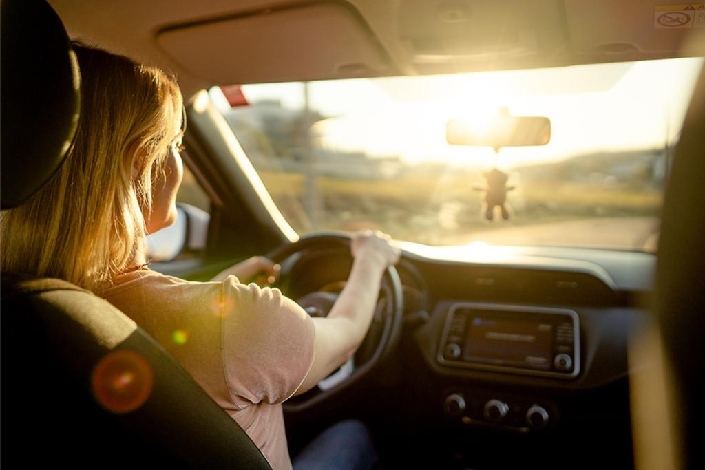 A woman drives into bright sunlight during golden hour, her face illuminated by glare coming through the windshield, illustrating the visual strain of low-angle winter sun in Colorado.