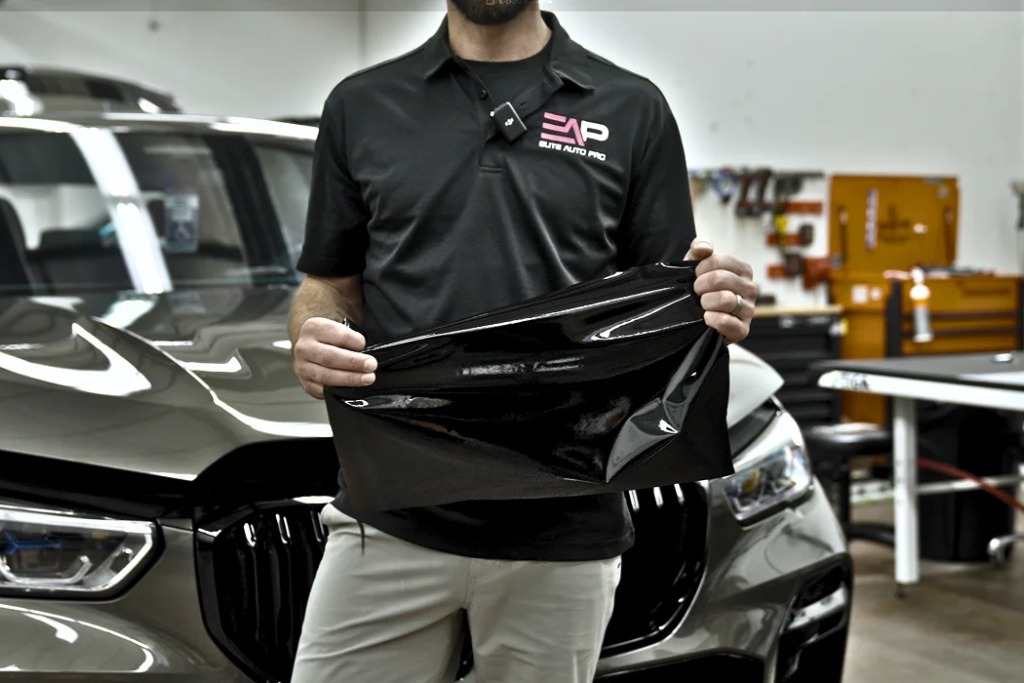 An Elite Auto Pro technician in a branded black polo shirt holds a sheet of Geoshield Pro Nano Ceramic IR window tint in front of a luxury SUV inside a professional garage setting.