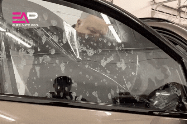 A technician at Elite Auto Pro carefully installs ceramic window tint on a car door, pressing out air and water bubbles during the application process inside a garage.
