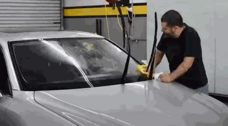 A technician in a detailing garage uses a cloth to apply and smooth out window tint across the full windshield of a silver car.