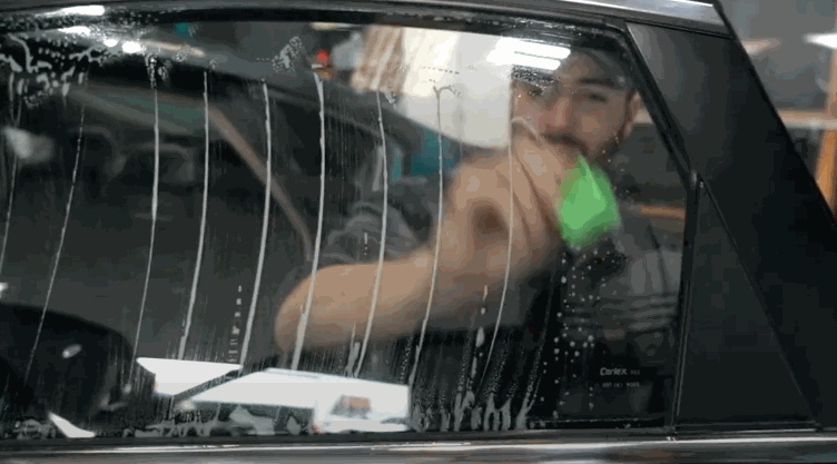Man applying window tint on a car's side window using a green squeegee, with streaks of soapy water visible on the glass.