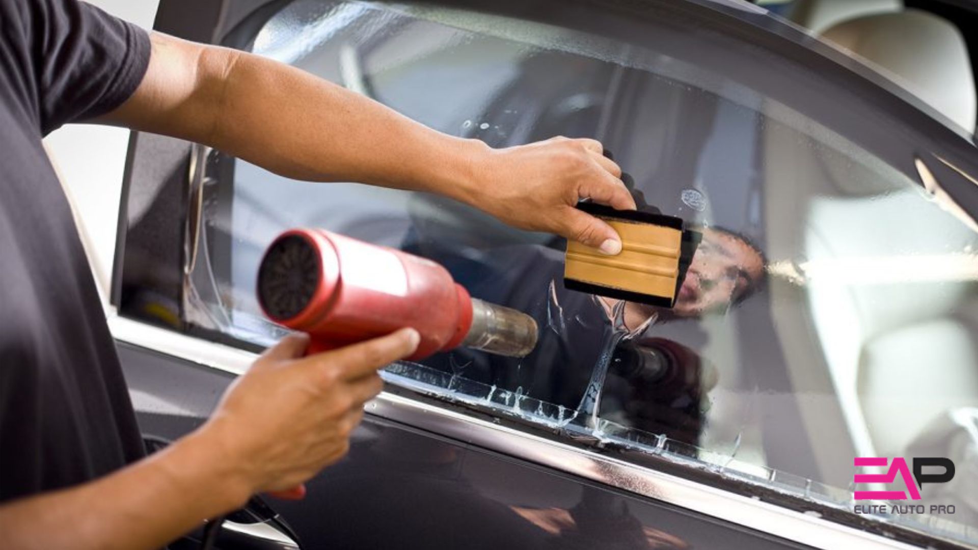 Person using a heat gun and a squeegee to shrink and adhere window tint film to a car window, with the Elite Auto Pro logo in the corner.